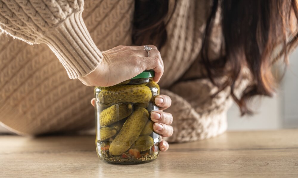 Frau mit Verlobungsring am Finger öffnet in beigen Pullover ein Glas mit Gewürzgurken. | © Getty Images / MarianVejcik
