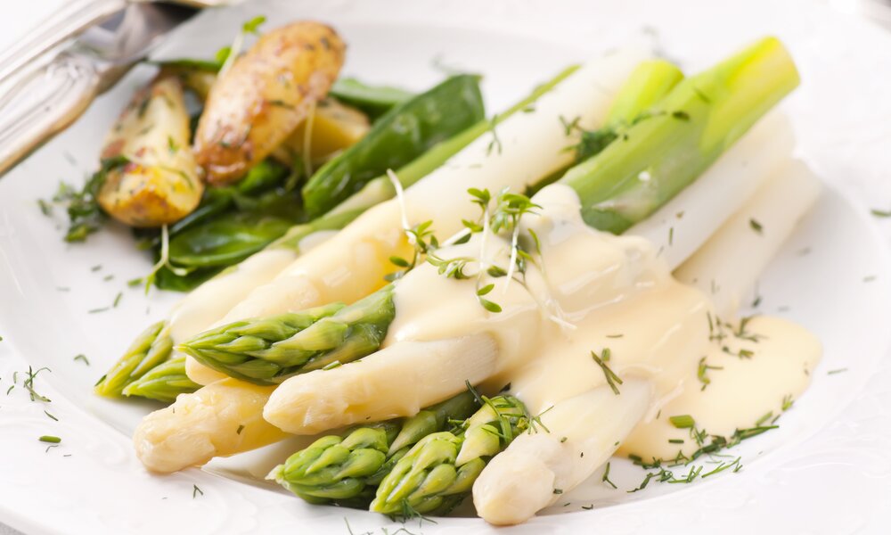 Spargel in Nahaufnahme begossen mit Sauce Hollandaise, angerichtet auf einem weißen Teller, daneben Kartoffeln. | © Getty Images / hlphoto