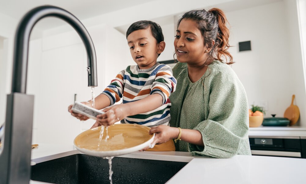 Eine junge Frau hält einen kleinen Jungen an ein Waschbecken und der Junge hält einen Teller unter laufendes Wasser. | © Getty Images / kate_sept2004