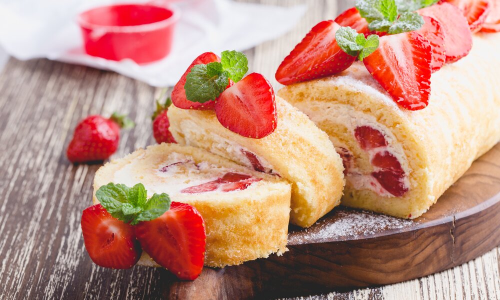 Aufgeschnittene Erdbeer-Biskuitrolle mit frischen Beeren und reichlich Puderzucker garniert. | © Getty Images / 	istetiana