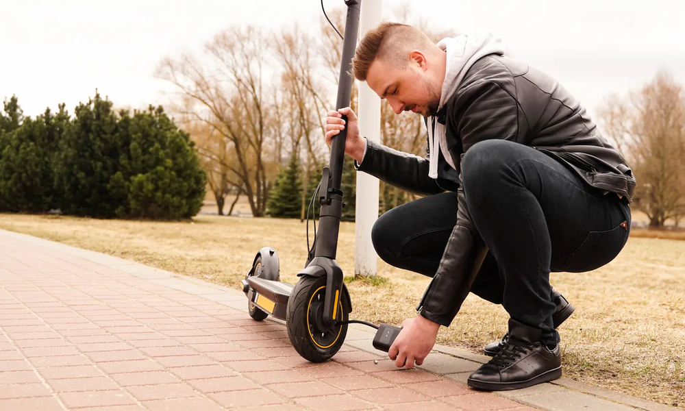 Junger Mann von der Seite in dunkler Lederjacke und Jeans kniet vor schwarzem E-Scooter und hält ihn mit einer Hand fest, mit der anderen Hand hält er eine kleine schwarze Akku-Luftpumpe an das Vorderrad, auf hellem Steinweg vor Grünfläche | © Adobe/ brillianata