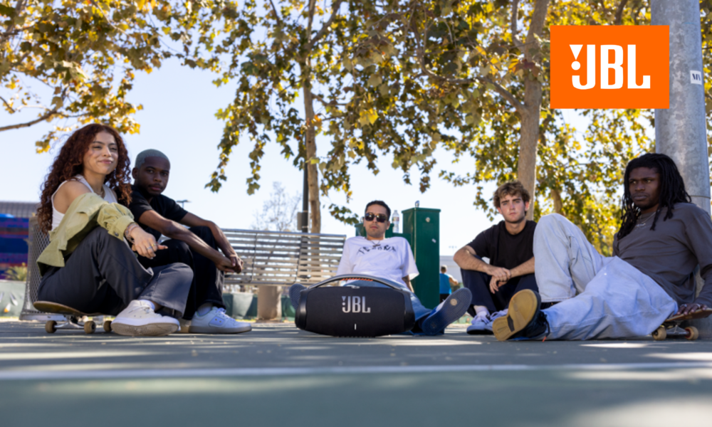 Junge Menschen sitzen rund um die JBL Boombox, dahinter ein Baum und eine Parkbank. | © JBL