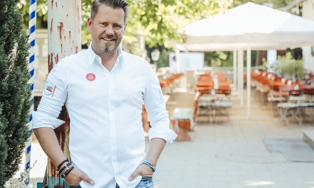 Mike Süsser in weißem Hemd und Jeans steht in einem sonnigen, leeren Biergarten. | © Kabel Eins