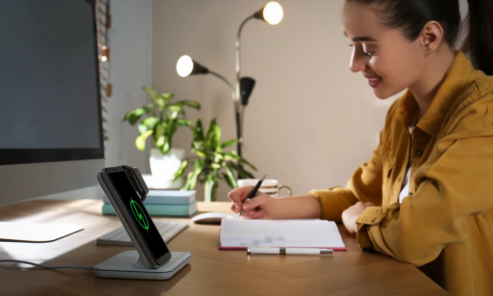 Junge Frau lächelnd von der Seite am Schreibtisch schreibt mit Stift in Notizbuch auf hellem Holztisch, während vor ihr ihr Smartphone auf einem Wireless Charger Ständer lädt und eine grüne Batterie anzeigt | © Adobe/ New Africa