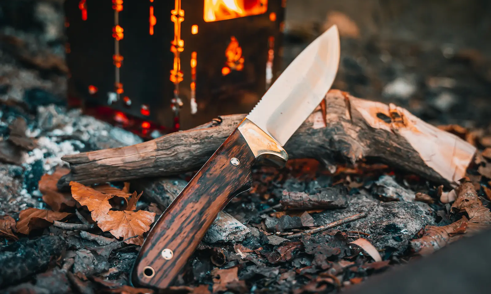 Outdoor-Messer mit Holzgriff und silberner Klinge an Aststück angelehnt auf rußigem Boden, dahinter verschwommen Metallkasten in dem Glut orange leuchtet | © Adobe/ Andre