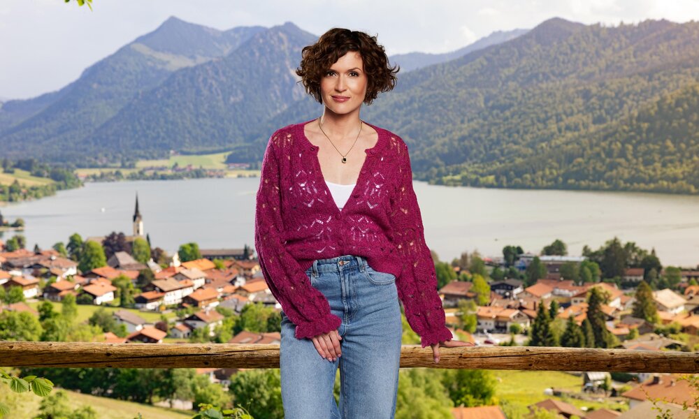 Eine braunhaarige Frau in lila Pullover und Jeans lehnt lächelnd an einem Holzgeländer mit Blick auf einen See und ein Dorf. | © Seven.One/Marc Rehbeck 