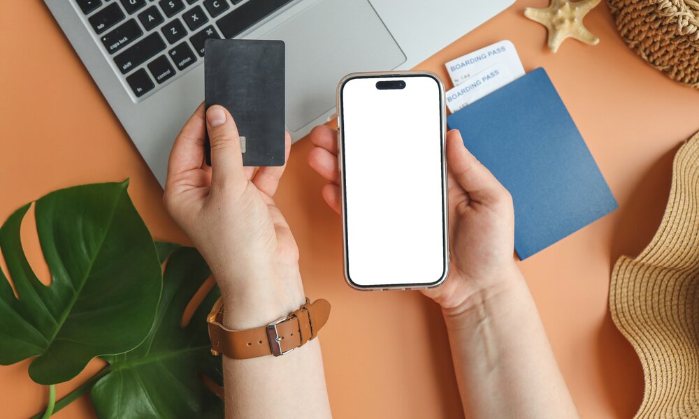 Zwei Hände halten ein Smartphone und eine schwarze Kreditkarte vor einem Laptop mit Reisepass und Boardingpässen. | © Getty Images/Bohdan Bevz
