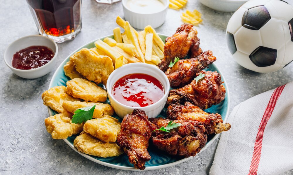 Gemischte Snackplatte mit Chicken Wings, Chicken Nuggets und Pommes, daneben ein Fußball, Dips und Getränke. | © Getty Images / Irina Kozmova