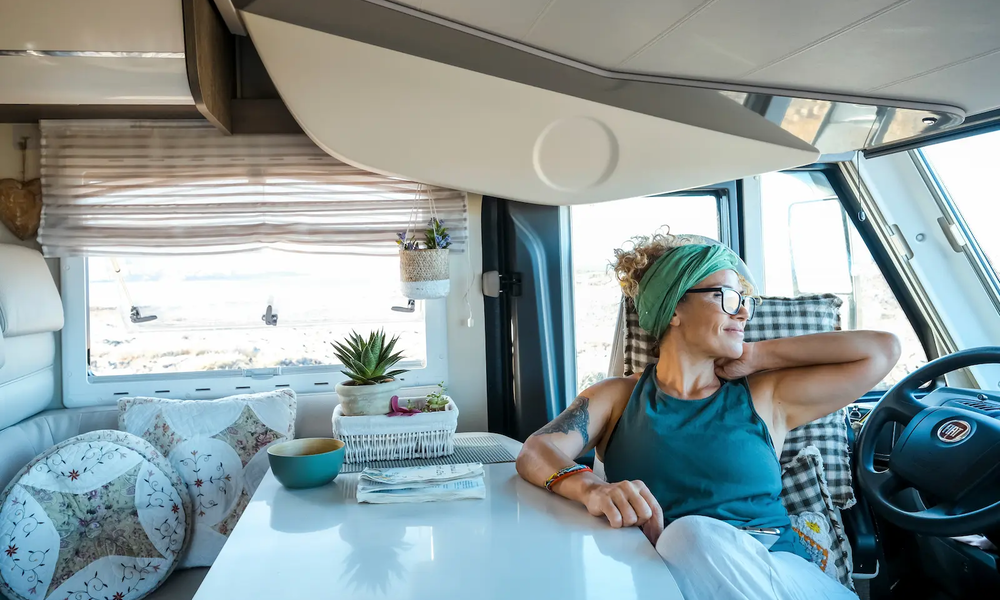 Lächelnde Frau mittleren Alters in Shirt mit Brille und hochgesteckten Haaren sitzt im Wohnmobil an Tisch, lässig zurückgelehnt und schaut aus Fenster | © Simona/ Adobe Stock