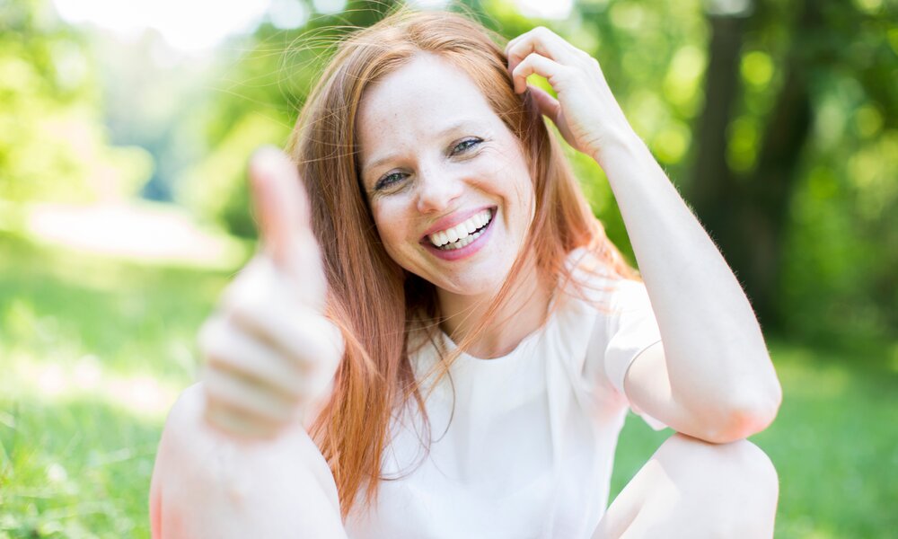 Lachende Frau mit roten Haaren im weißen Shirt gibt im sonnigen Park ein Daumen-hoch-Zeichen. | © Adobe Stock/ drubig-photo