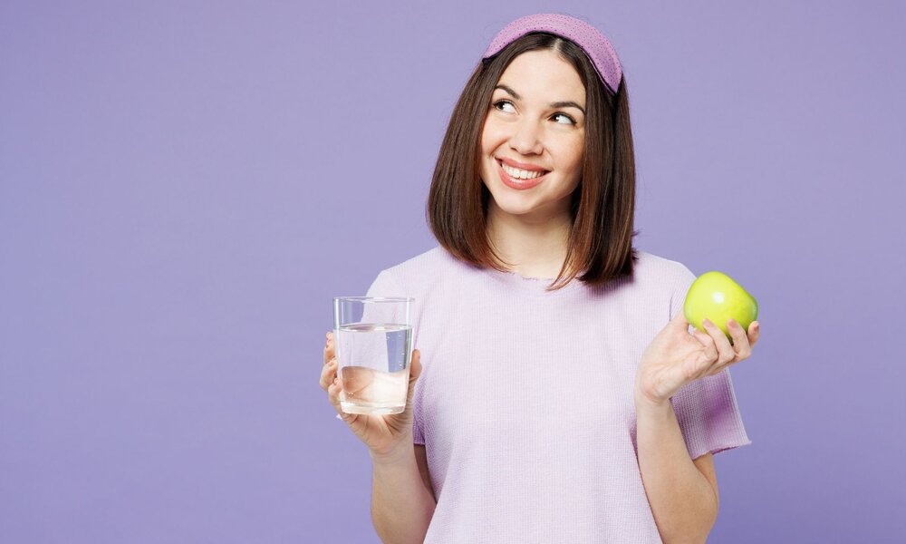 Eine junge Frau mit braunen Haaren und lila Stirnband hält lächelnd ein Glas Wasser und einen grünen Apfel vor lila Hintergrund. | © Adobe Stock/ ViDi Studio