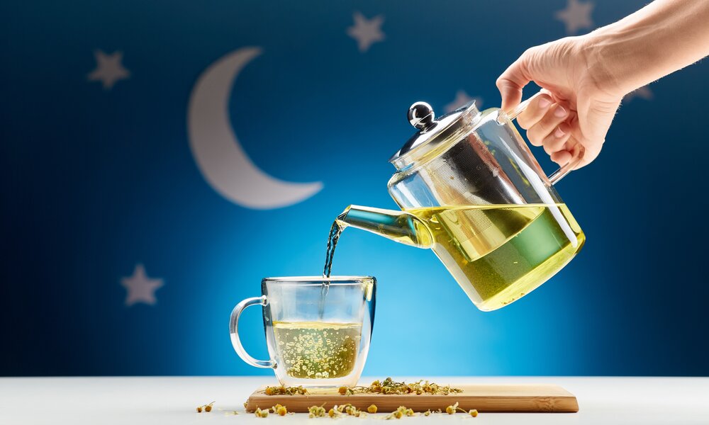 Hand gießt heißen Kamillentee aus einer Glaskanne in eine Tasse vor blauem Hintergrund mit Mond und Sternen. | © Adobe Stock/ Syda Productions