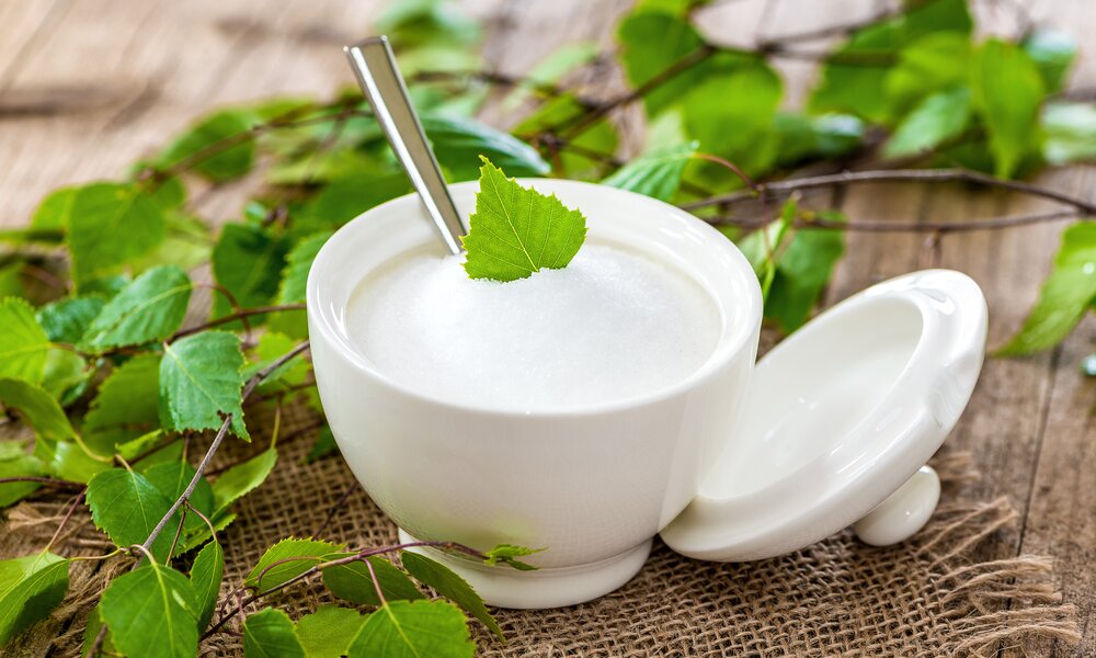 Zuckerdose aus Porzellan gefüllt mit Xylit, verziert mit einem grünen Blatt, umgeben von grünen Zweigen auf Jutegewebe. | © Adobe Stock/ Lumixera