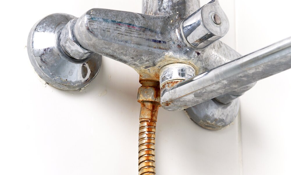 Nahaufnahme eines verrosteten Duschschlauchs an einem verkalkten Wasserhahn vor weißer Wand. | © Adobe Stock/ ZUMRUT