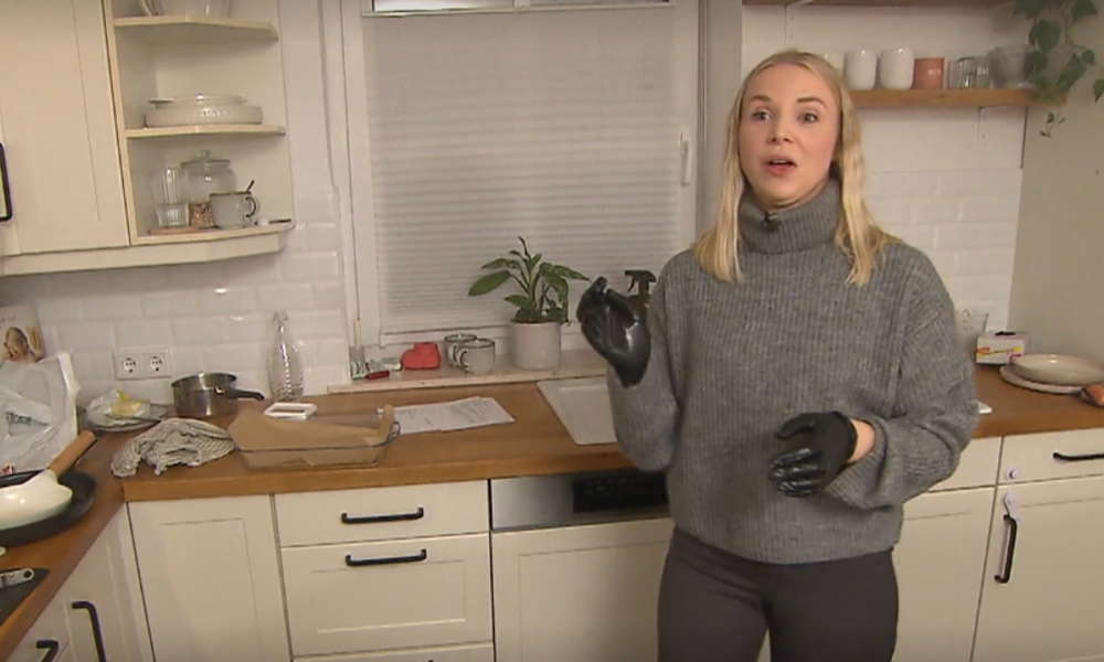 Eine junge Frau mit blonden Haaren steht in einer modernen Küche, trägt einen grauen Pullover und schwarze Handschuhe. | © Screenshot Vox/RTL+ 