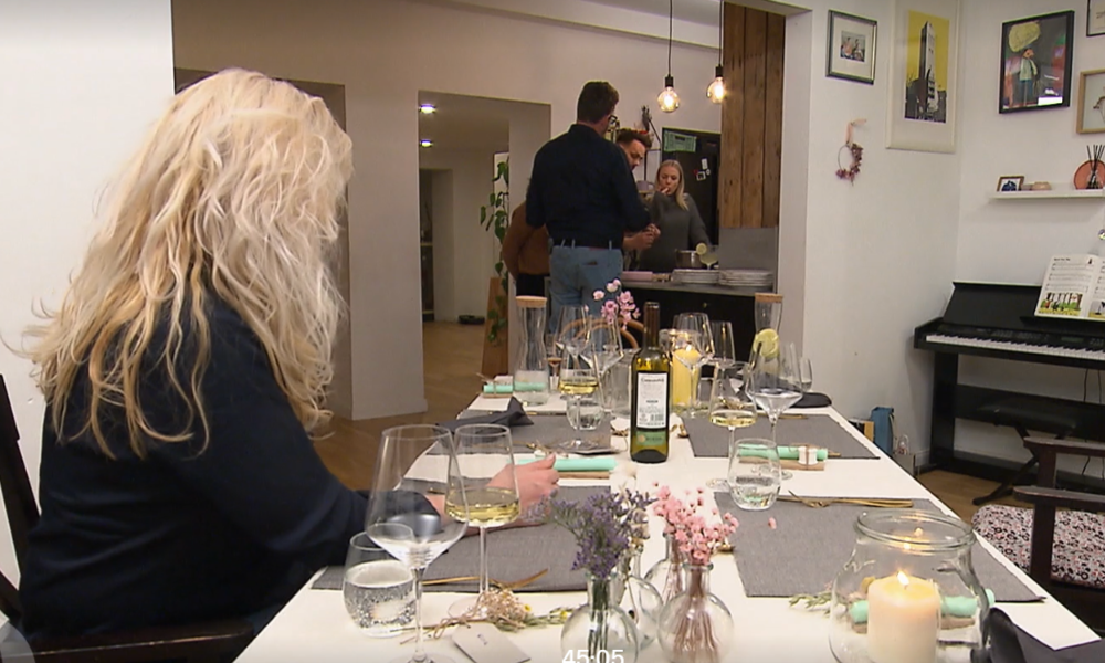 Eine blonde Frau sitzt an einem gedeckten Tisch, während im Hintergrund drei Personen in einer modernen Küche stehen. | © Screenshot Vox/RTL+ 