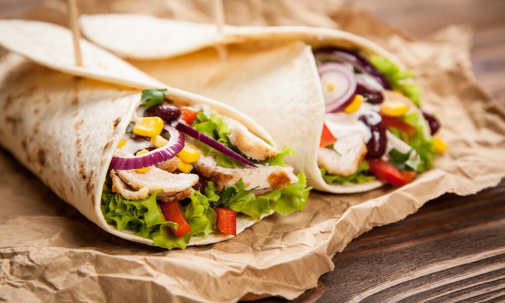 Zwei Wraps mit Hähnchen und Salatfüllung liegen auf Backpapier auf einem Holzuntergrund. | © Adobe Stock / George Dolgikh