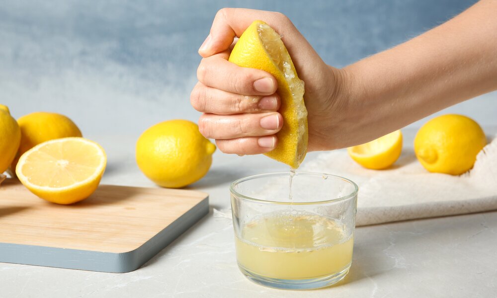 Eine Hand presst eine halbe Zitrone aus, während der Saft in ein Glas tropft, daneben liegen ganze und halbe Zitronen auf einem Schneidebrett. | © Adobe Stock/ New Africa