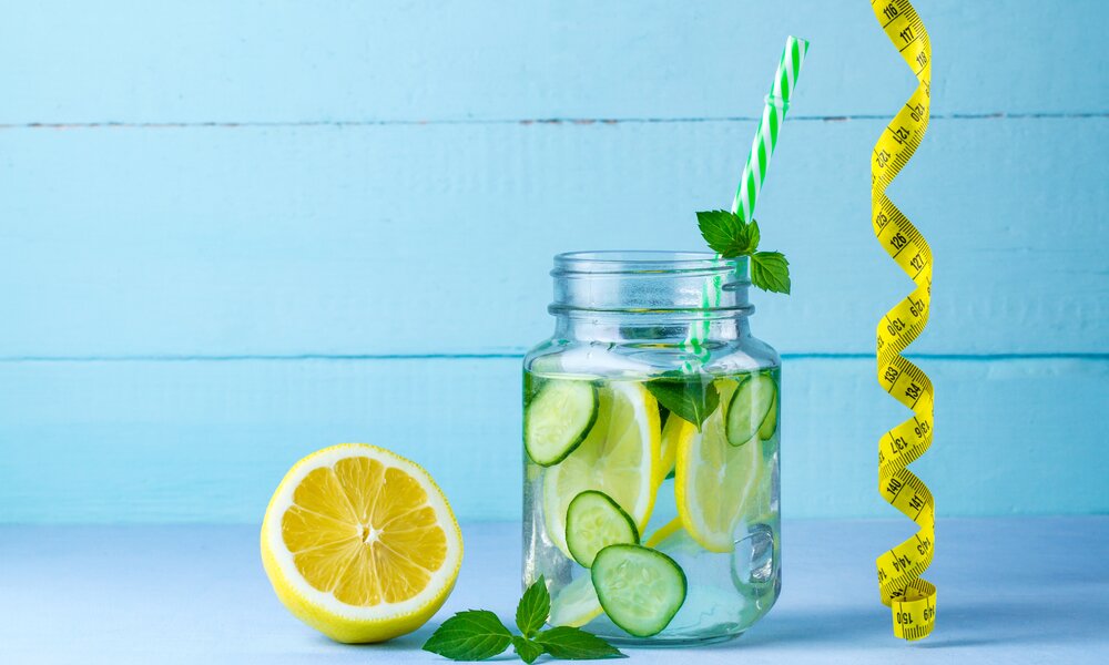 Ein Glas Wasser mit Gurkenscheiben, Zitronenscheiben und Minzblättern, daneben eine halbe Zitrone und ein gelbes Maßband auf hellblauem Hintergrund. | © Adobe Stock/ Goffkein
