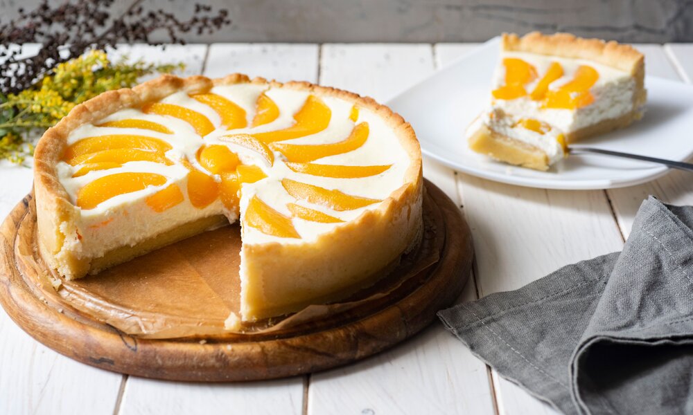 Angeschnittener Joghurtkuchen mit Pfirsich, auf einem Teller angerichtet, dahinter das Stück Kuchen auf einem anderen Teller. | © Adobe Stock /  Светлана Терлюкевич