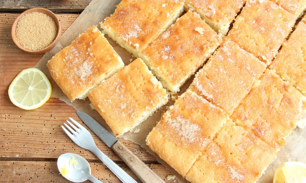 In Stücke geschnittener Zitronenkuchen vom Blech, daneben Messer und gaben und frische Zitronenscheiben. | © Adobe Stock / elfstudioart