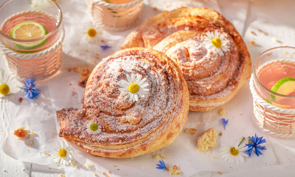 Zwei mit Puderzucker bestreute Hefeschnecken in sommerlicher Umgebung, daneben Limonaden in Gläsern. | © Adobe Stock / shaiith