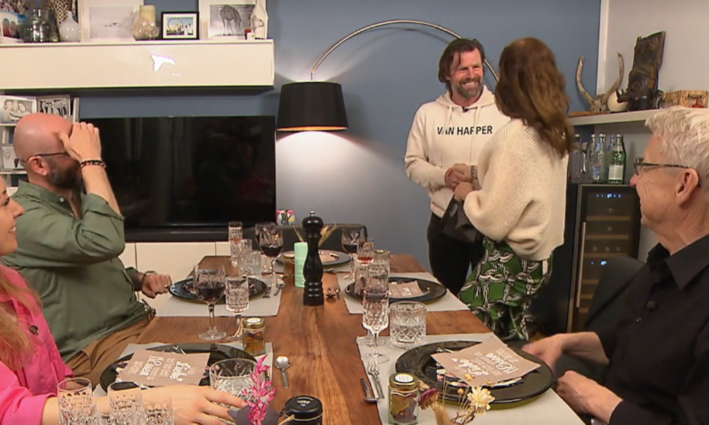 Ein Mann im weißen Hoodie schüttelt einer Frau in grünem Rock die Hand, während drei andere Personen am Esstisch sitzen und lachen. | © Screenshot Vox/RTL+ 