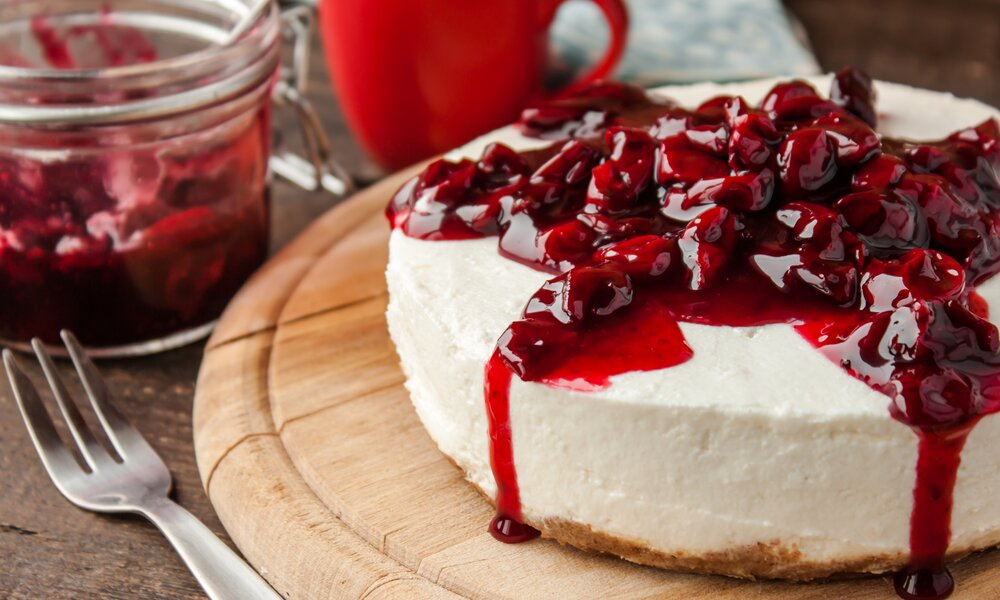 Cremiger Joghurtkuchen mit einem Kirschkompott-Topping, daneben Gabel, Kaffeetasse und mehr Kompott in Glas. | © Adobe Stock / yuliiaholovchenko