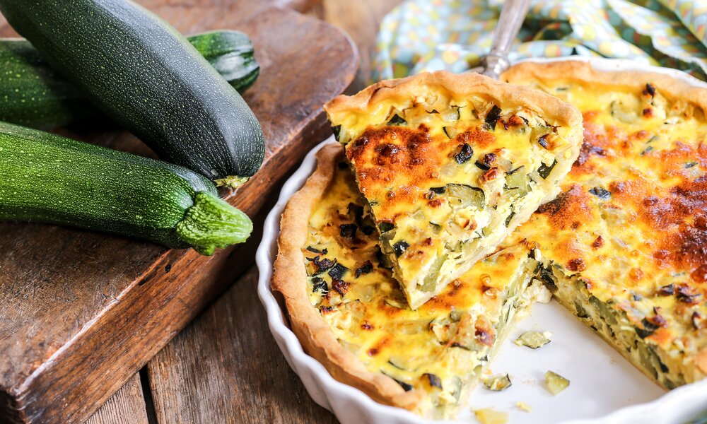 Angeschnittener Zucchini-Zwiebelkuchen in einer Tarte-Form, daneben frische Zucchini auf einem Holzbrett. | © Adobe Stock / Jérôme Rommé