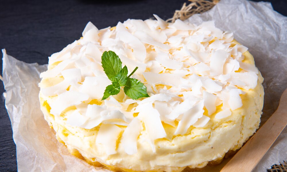 Luftiger Kokos-Quarkkuchen auf etwas Backpapier, daneben Kokosraspeln in einer Kokosnuss. | © Adobe Stock / Dar1930