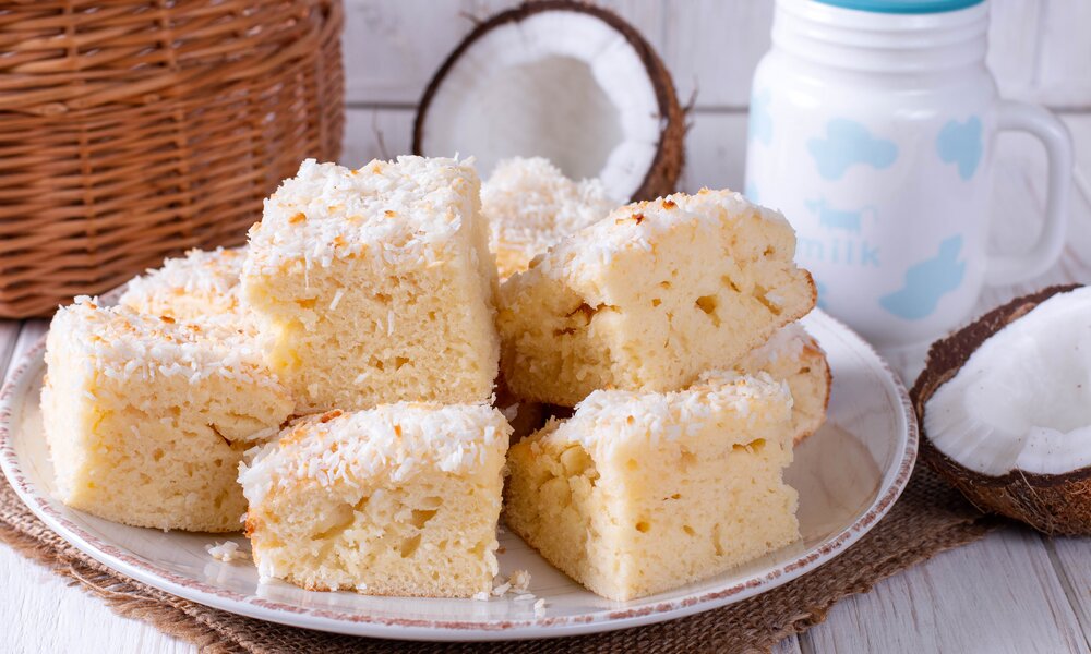 Kleine Kokos-Quarkkuchen-Stücke auf einem Teller drapiert, dahinter eine offene Kokosnuss und eine Milchkanne. | © Adobe Stock / qwartm