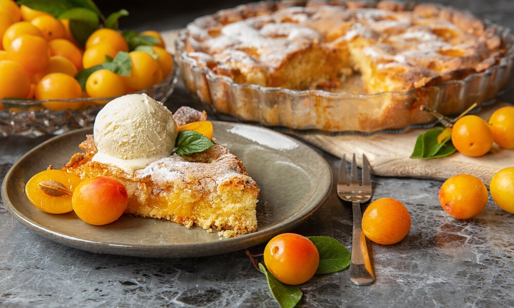 Stück Mirabellen-Kuchen mit Eis auf einem Teller, dahinter der ganze Kuchen und frische Mirabellen.  | © Adobe Stock / Elena