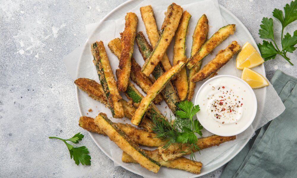 Mehrere Zucchini-Pommes auf einem weißen Teller angerichtet und daneben in einer weißen Schale befindet sich ein Joghurtdip. | © Adobe Stock / anna_shepulova
