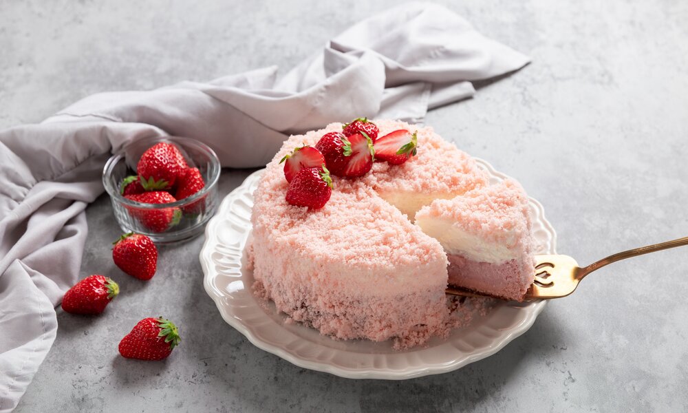 Angeschnittener Kokos-Quarkkuchen mit frischen Erdbeeren, angerichtet auf einem weißen Teller, daneben weitere frische Erdbeeren und ein Tuch. | © Getty Images / yumehana
