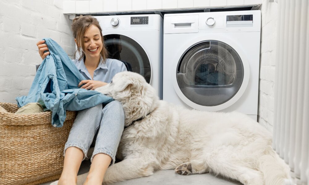 Junge Frau sitzt lächelnd auf dem Boden vor Waschmaschine, hält ein blaues Hemd und wird von großem, weißen Hund liebevoll angestupst. | © Adobe Stock/ rh2010