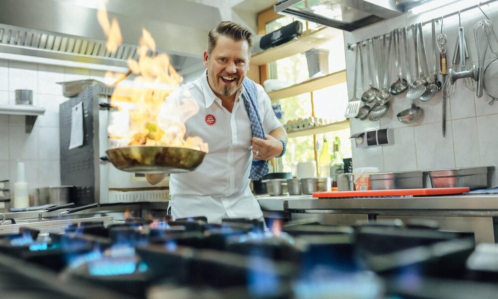 Mike Süsser trägt eine weiße Uniform und schwenkt eine brennende Pfanne über einem Gasherd in einer modernen Küche. | © Kabel Eins