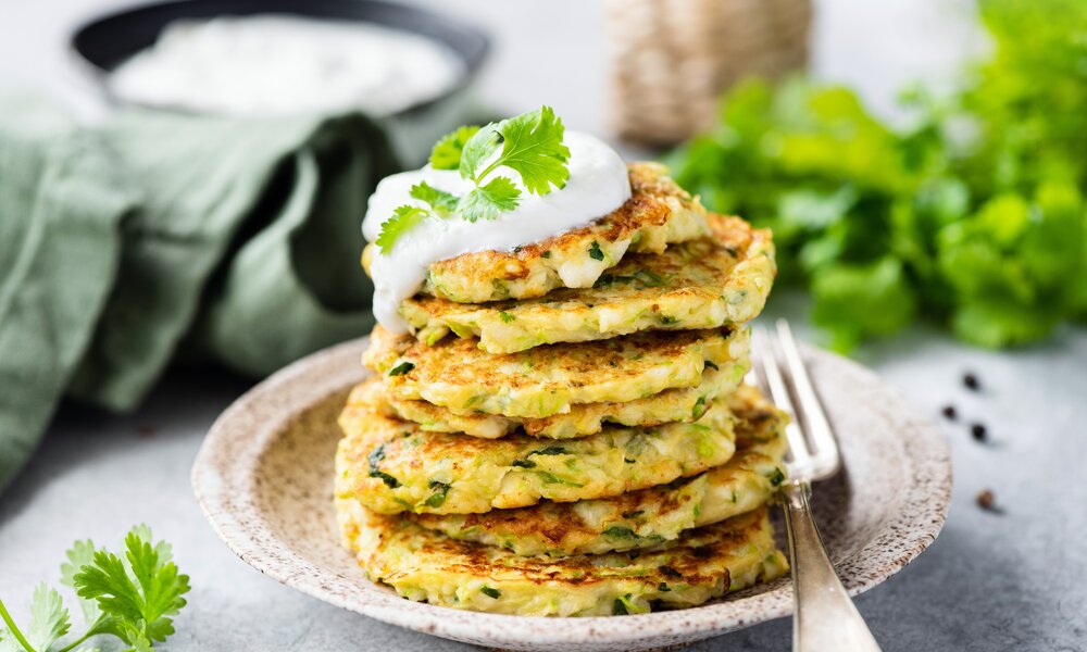 Gestapelte Zucchini-Pfannkuchen auf einem Teller, dahinter etwas Quark, und frische Petersilie. | © Adobe Stock / Vladislav Noseek