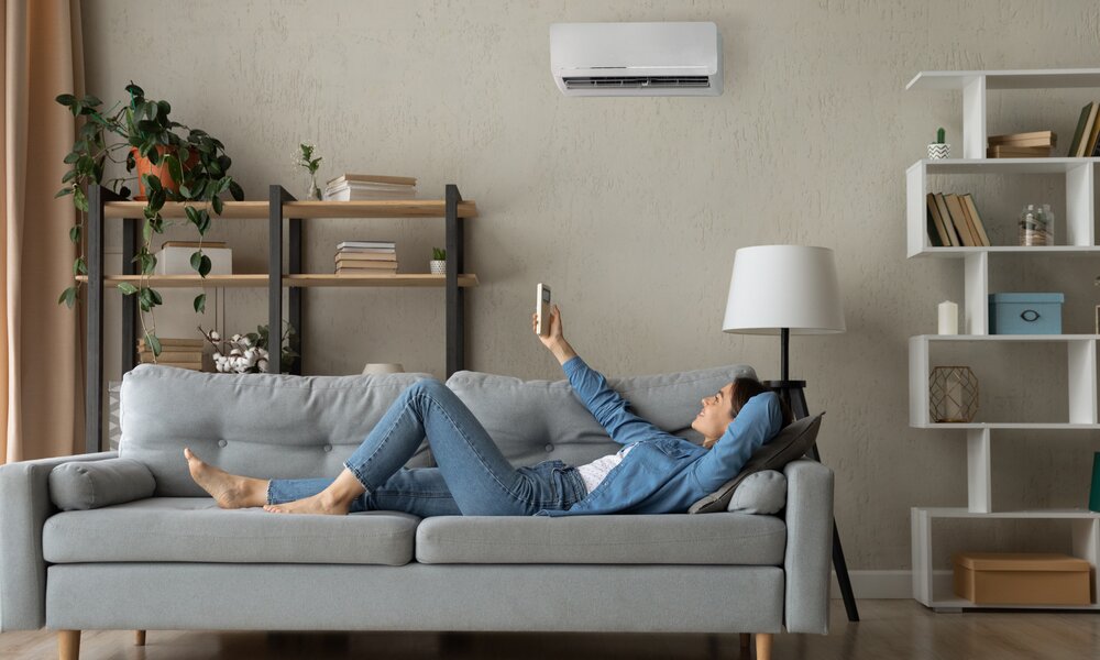 Frau liegt auf Sofa und schaltet Klimaanlage ein | © Getty Images / fizkes