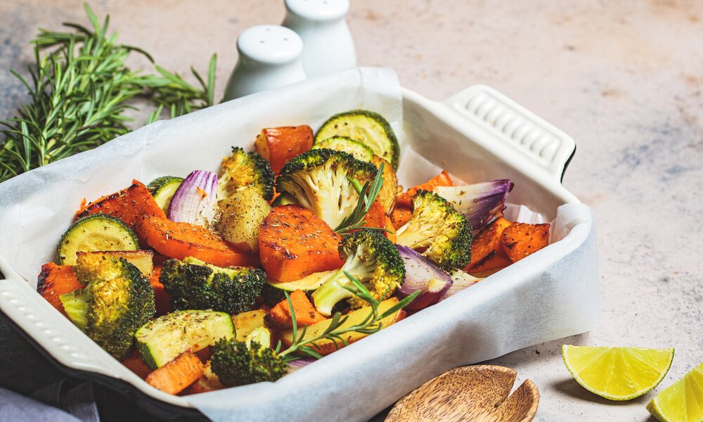 Brokkoli, Zucchini und Möhren befinden sich zusammen mit etwas Oregano und einem Rosmarinzweig in einer Auflaufform. Das Gemüse kommt gerade frisch gebraten aus dem Ofen und steht auf dem Tisch neben einen Salz und Pfefferstreuer. Daneben liegt eine Zitronenspalte. | © Getty Images / vaaseenaa