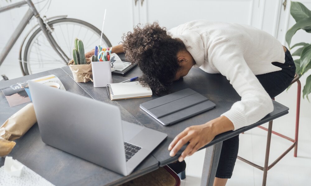 Frau mit Zopf und in legerer Kleidung sitzt auf einem Stuhl und liegt mit dem Kopf auf dem Schreibtisch. Vor ihr steht ein geöffneter Laptop, ein Block, Stifte und ein kleiner Kaktus. Im Hintergrund steht ein Fahrrad und eine Pflanze. | © Getty Images / Westend61