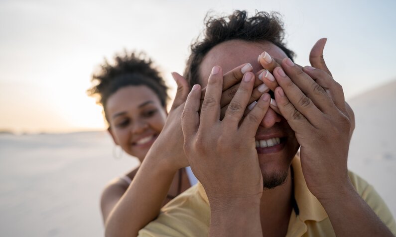 Eine Frau hält einem Mann die Augen zu. Sie befinden sich in einer Strand-Kulisse. | ©  Getty Images / andresr