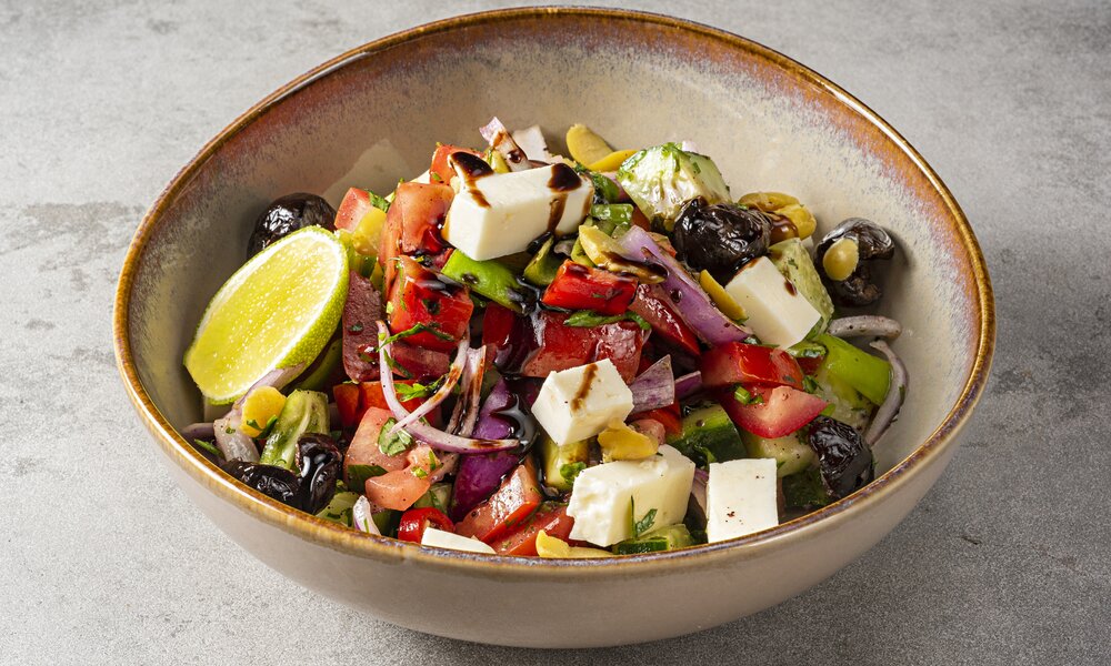 Ein Salat in einer beigen Schüssel angerichtet, in ihm enthalten sind Feta, Oliven, Salat, Zitrone, Gurke und Tomaten. Die Schüssel steht auf einem grauen Untergrund, der Salat ist mit einem dunklen Essig-Dressing beträufelt. | © Getty Images / BURCU ATALAY TANKUT