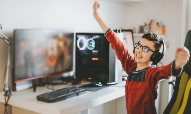 Junge freut sich über Erfolge beim Computerspielen, reißt die Arme hoch, während er am Schreibtisch vor seinem PC sitzt. | © Getty Images/Carol Yepes