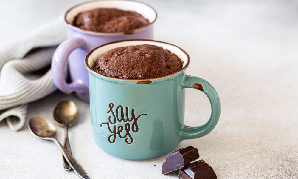 Zwei Tassen mit fertigem Schoko-Küchlein. Eine Tasse ist blau, die andere lila. Auf den Tassen steht "say yes" und neben den Tasser liegen zwei Teelöffel, zwei Stücke Schokolade und ein Geschirrtuch. | © Getty Images / Iryna Melnyk