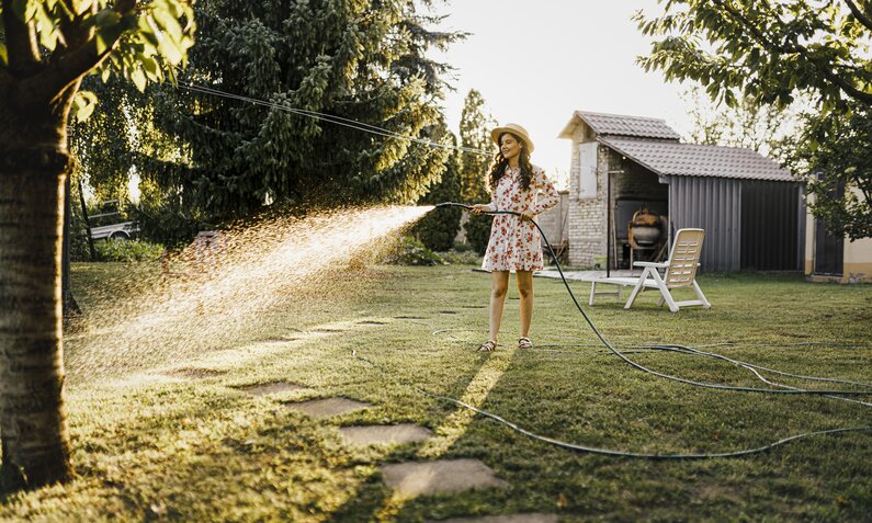 Frau in Sommerkleid und Hut bewässert ihren Rasen mit einem Schlauch  | ©  Getty Images / Eleganza