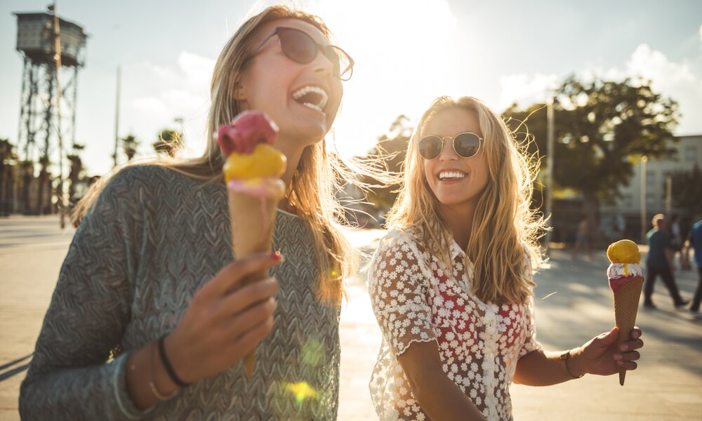 Zwei junge Frauen in Nahaufnahme lachen sich an und tragen Sonnenbrillen. Jede hält ein Eis in der Hand. Es scheint die Sonne und es herrscht ein sommerliches Ambiente.  | © Getty Images / Georgijevic