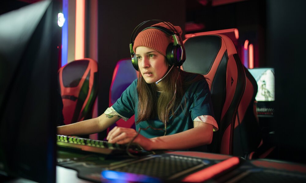 Eine junge Frau mit Mütze auf dem Kopf spielt konzentriert an einem PC. Sie hat ein Headset auf dem Kopf und sitzt in einem speziellen Gaming-Stuhl. | © Getty Images/ Marko Geber