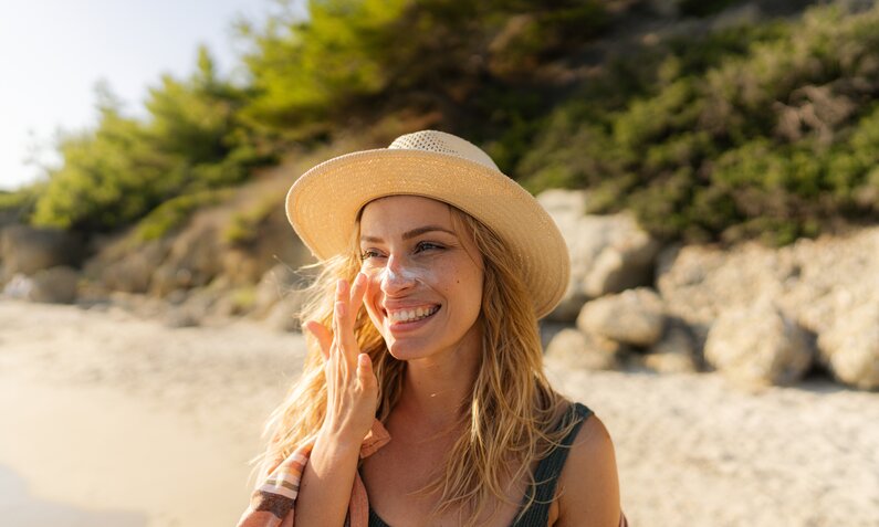 Blonde und junge Frau trägt einen Hut und ist am Strand. Sie trägt sich Streifen von Sonnencreme auf die Wangen und lächelt dabei. | © Getty Images / AleksandarNakic