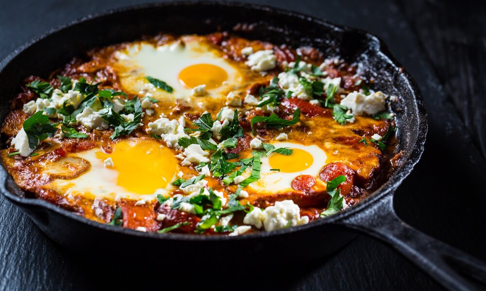 Shakshuka in einer schwarzen, gusseisernen Pfanne in Nahaufnahme. Zwei Eier sind in der Mitte des Gerichts und ein schwarzer Holztisch als Untergrund. | © Getty Images / brebca