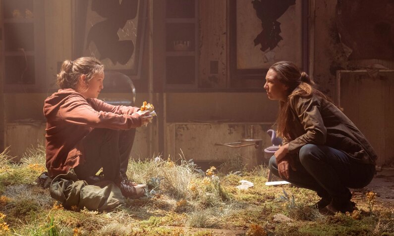 Ellie-Darstellerin Belly Ramsay sitzt in einem Lichtstrahl auf dem Boden eines Raums, der mit Grün bewachsen ist. Tess-Darstellerin Anna Torv hockt rechts von ihr und spricht mir ihr. | © HBO
